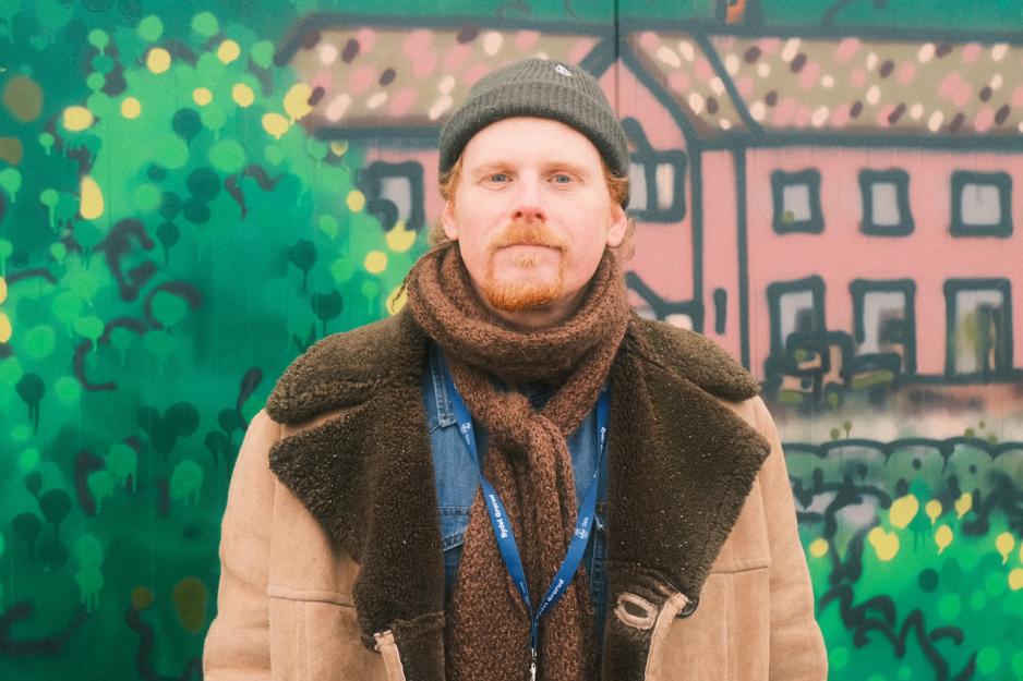 Mann med skjerf, lue og k&aring;pe foran et veggmaleri