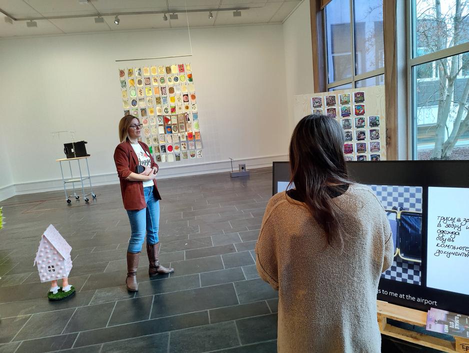 Kunstgalleri. Dame i midten med olabukse og r&oslash;d blazer. Ser ryggen til dame med langt brunt h&aring;r. 