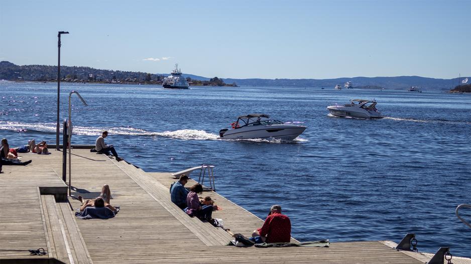 Flere mennesker sitter og soler seg p&aring; en brygge ved havet. To b&aring;ter seiler mot hverandre.
