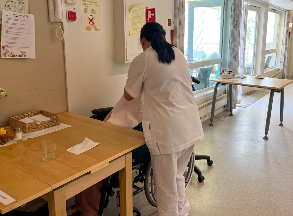 Helsefagarbeider st&aring;r med ryggen til og gir en eldre person en kopp med noe &aring; drikke.