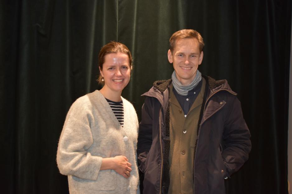 En dame med brunt oppsatt h&aring;r, stripete genser og beige cardigan holder seg p&aring; magen. Ektemann ved siden av. Han har tre jakker p&aring; seg. Og en hals i halsen. 