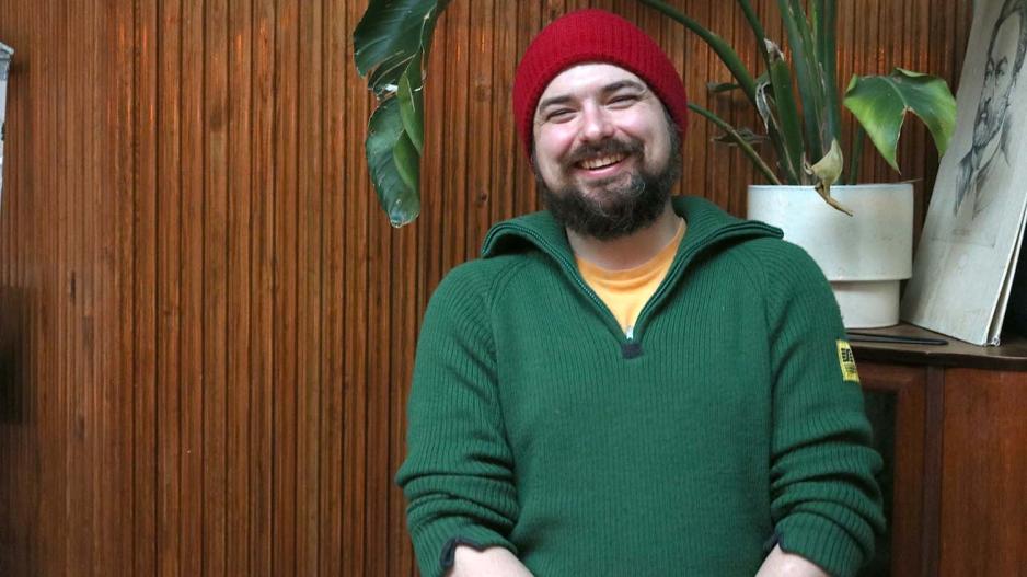 Portrett av Daniel Kosacki. Han st&aring;r foran en brun trevegg og har en plante bak seg.