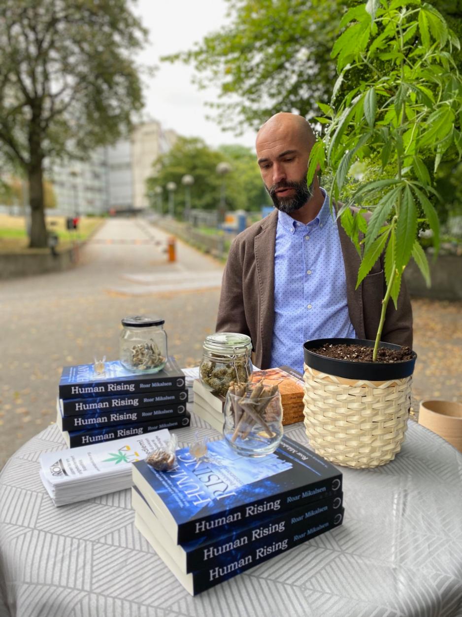 Roar Mikalsen st&aring;r ved bord med krukker med stoff, b&oslash;ker og en cannabisplante. 