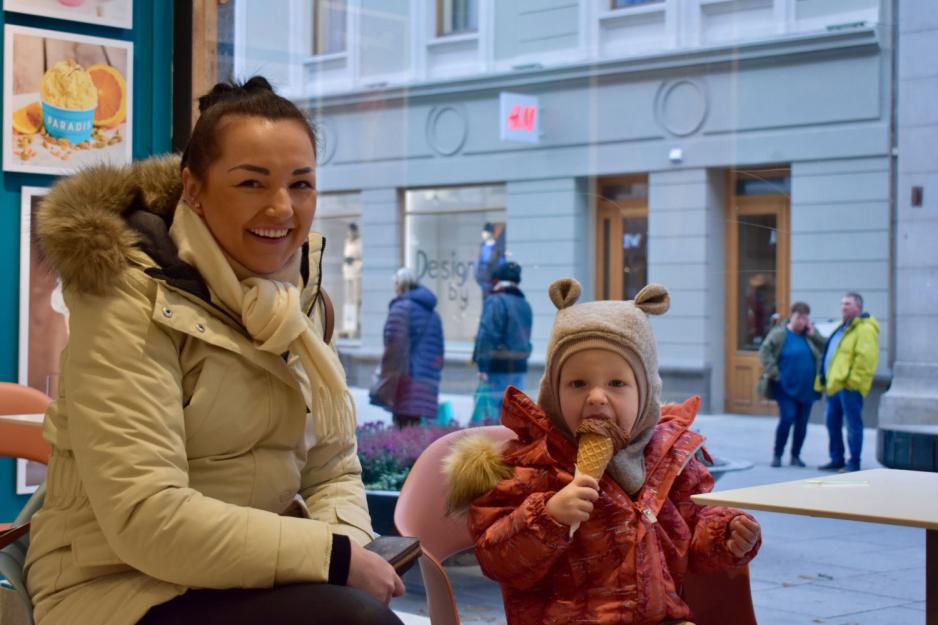 Liten gutt med bamselue og orange jakke spiser is, mens moren, en dame med m&oslash;rkt h&aring;r og lyst skjerf, sitter p&aring; huk ved siden av