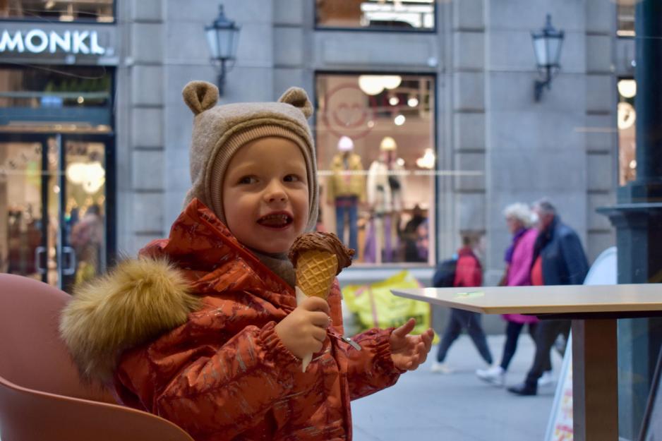 Liten gutt med bamselue og orange vinterakke spiser is p&aring; en isbar i Karl Johans gate. Utenfor er gaten travel. 