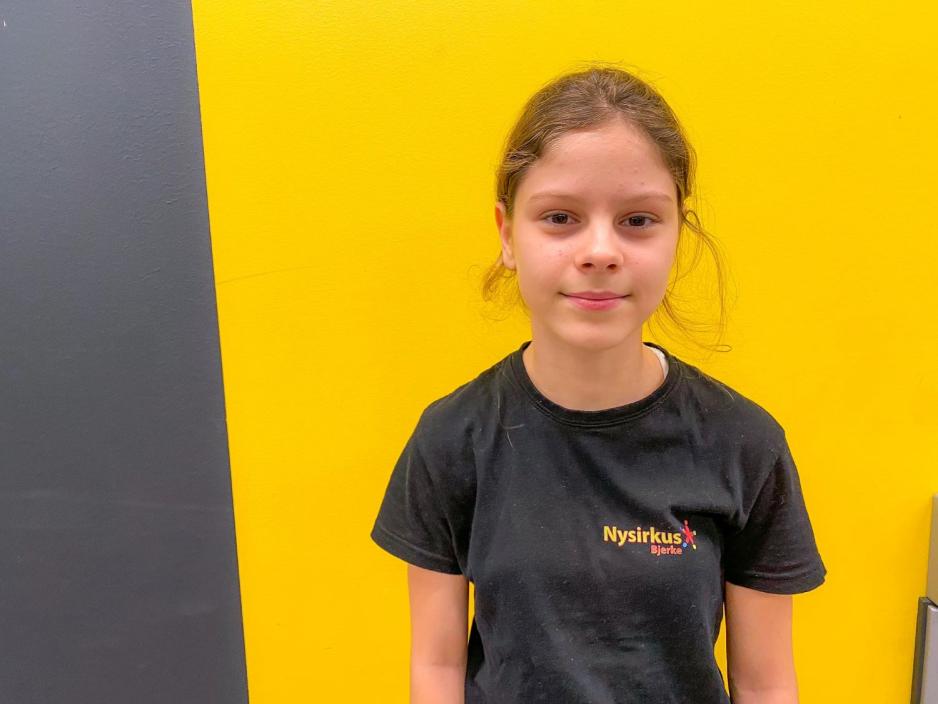 Ei smilende jente p&aring; 10 &aring;r som smiler. Hun har p&aring; seg en svart t-skjorte og st&aring;r foran et gul vegg. 