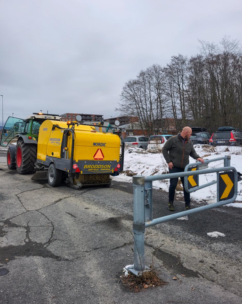 I bakgrunnen ser man en gr&oslash;nn traktor med gult feieslep, og i forgrunnen &aring;pner sj&aring;f&oslash;ren en bom som han skal kj&oslash;re gjennom.