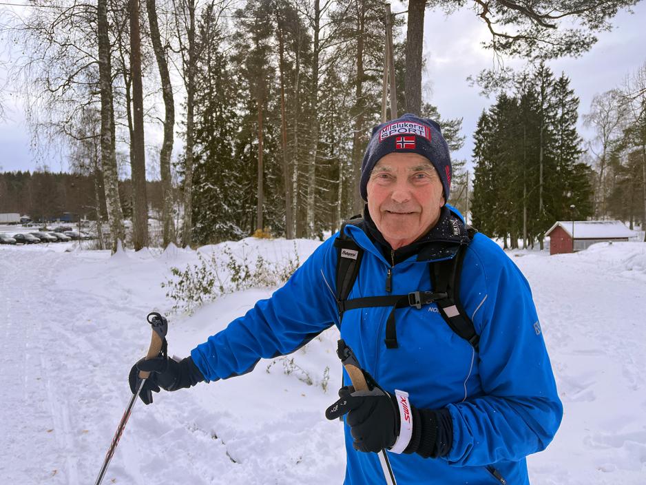 Eldre mann med med en m&oslash;rk lue, en bl&aring; jakke, sekk p&aring; ryggen og har p&aring; skiutstyr