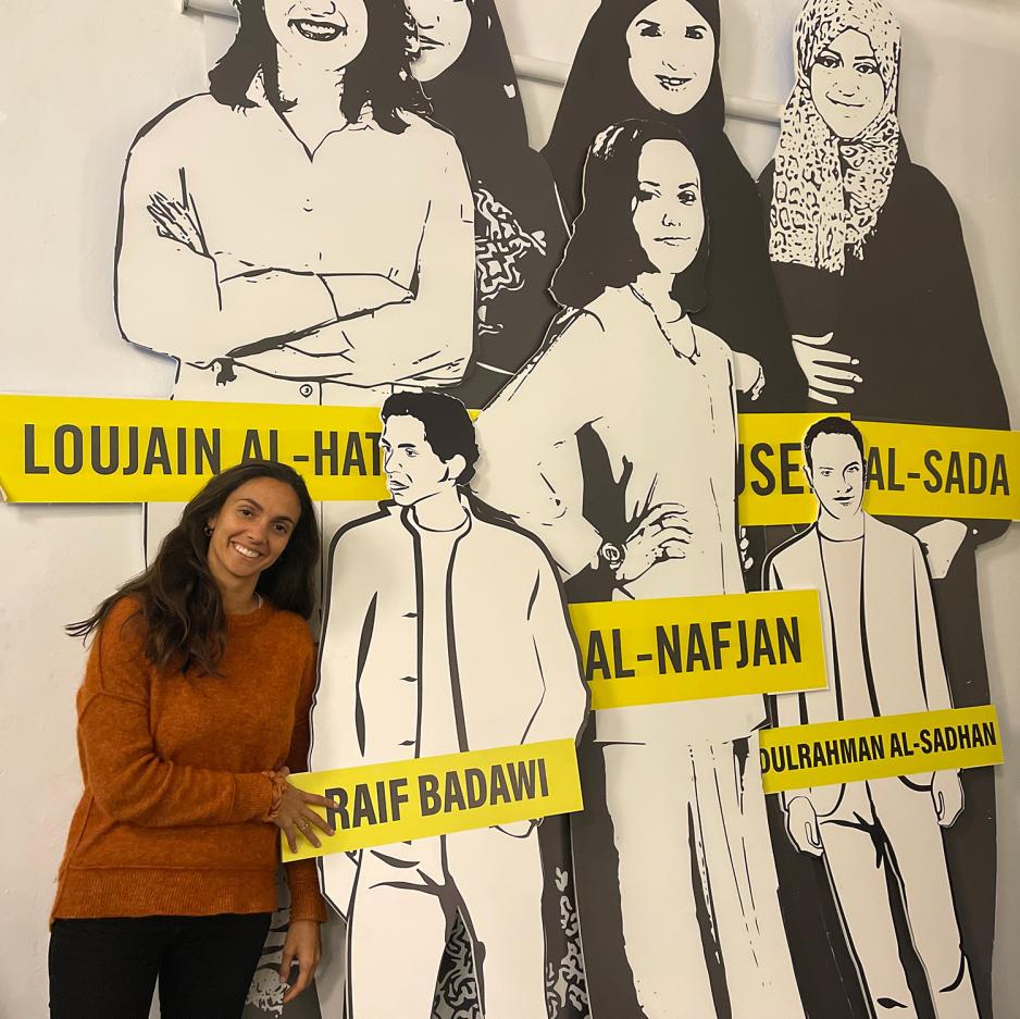 Ida Hellum st&aring;r foran en plakat av ulike menneskerettighets-aktivister i svar hvitt bilde. Hun har p&aring; seg en orange genser. 