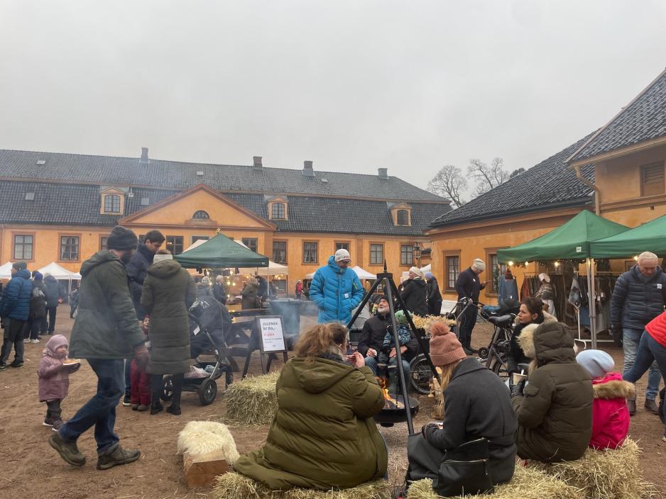 Bildet viser en gjeng som sitter og spiser mat p&aring; h&oslash;yballer rundt en b&aring;lpanne. I bakgrunnen ser man den oransje hovedbygningen p&aring; Bogstad G&aring;rd. 