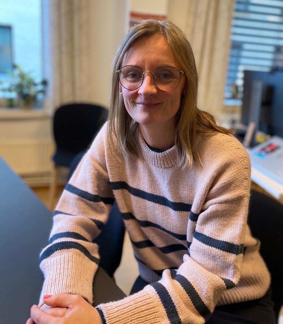 Et portrettbilde av Hege Undem Store p&aring; kontoret sitt. Hun har hendene samlet foran seg, og smiler til kameraet.