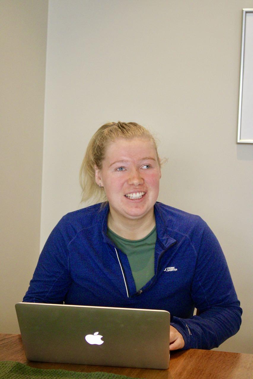 Portrett av Kristin Hauglid. Hun sitter med PC'en foran seg p&aring; spisebordet, og smiler.