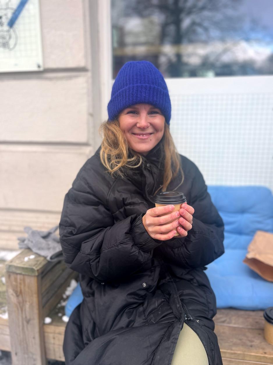 Bilde av en kvinne med en kaffekopp som sitter ute med en bl&aring; lue foran en kaf&eacute; 