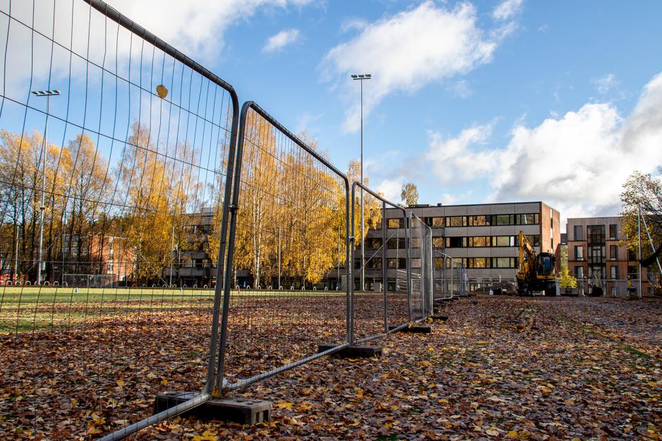 Kunstgressbane med mye l&oslash;v og et byggegjerdet med en gravemaskin og et boligbygg i bakgrunnen