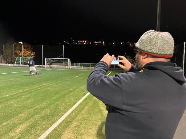Marius Helg&aring; bruker mobilen til &aring; filme en hendelse p&aring; fotballbanen i bakgrunnen.