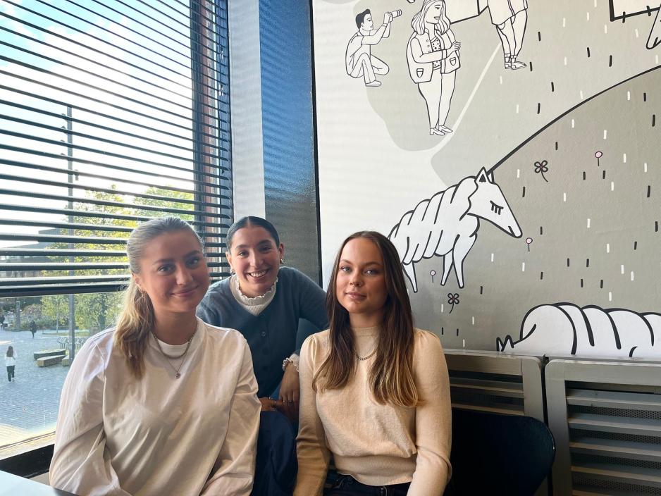 Tre jenter sitter ved et bord og smiler mot kamera