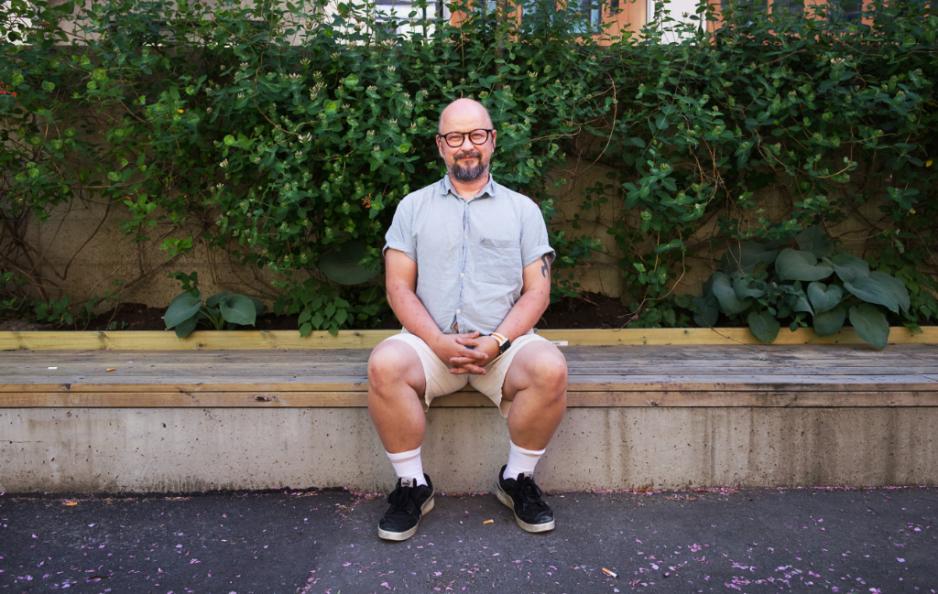 Portrett av Mikael Scott Bjerkeli hvor han sitter p&aring; en benk foran en busk. Hendene er foldet i fanget. 
