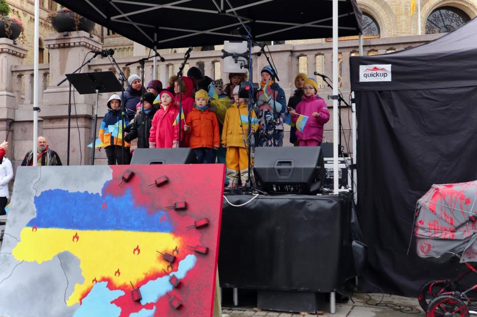 mange barn p&aring; en scene som holder det ukrainske flagget. de st&aring;r foran stortinget. foran dem er et maleri av Ukraina og Russland, der russland er r&oslash;dt og invaderer Ukraina.