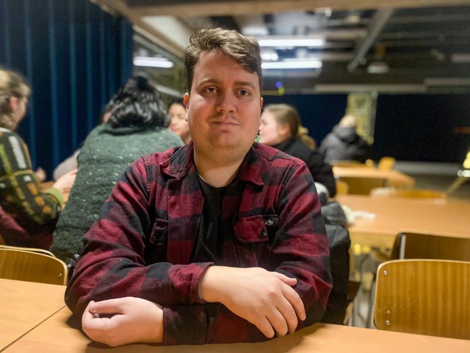 En mann som sitter ned ved et bord. Han har p&aring; seg en skjorte svart og r&oslash;dt. 