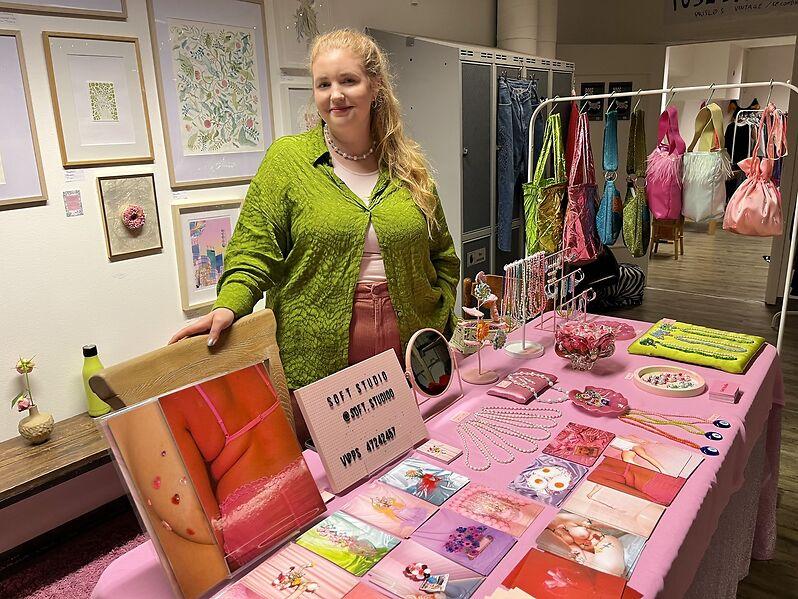 Fotograf og designer Sara Tonning bak sin markedsbod dekket med rosa duk, med postkort, smykker og et stativ med egenproduserte h&aring;ndvesker i sterke i&oslash;yenfallende farger.