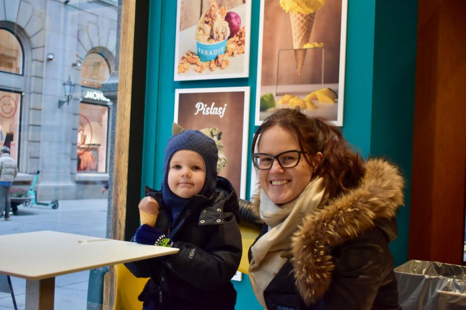 Glad dame med svarte briller og m&oslash;rkt h&aring;r sitter ved siden av s&oslash;nnen sin p&aring; omtrent to &aring;r. Han har m&oslash;rke kl&aelig;r og spiser p&aring; en kule sjokoladeis i kjeks. 