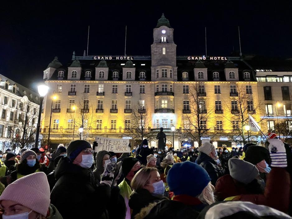 En godt p&aring;kledd folkemengde st&aring;r tett i tett med munnbind p&aring;. Noen av demonstrantene hever et banner det st&aring;r "lite lys og lite varme" p&aring;. 