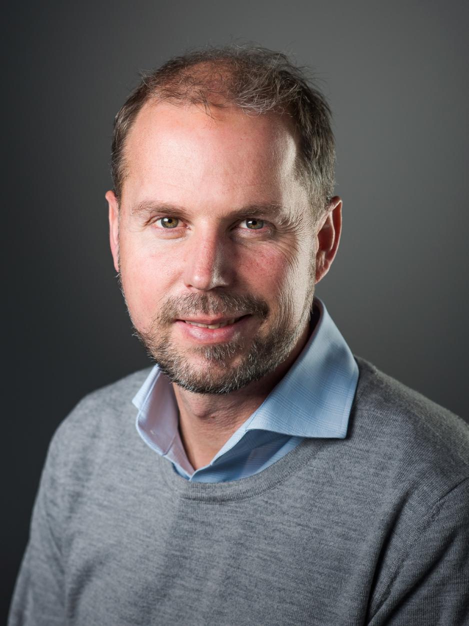 Mann med skjegg og brunt h&aring;r, smilende i en gr&aring; genser