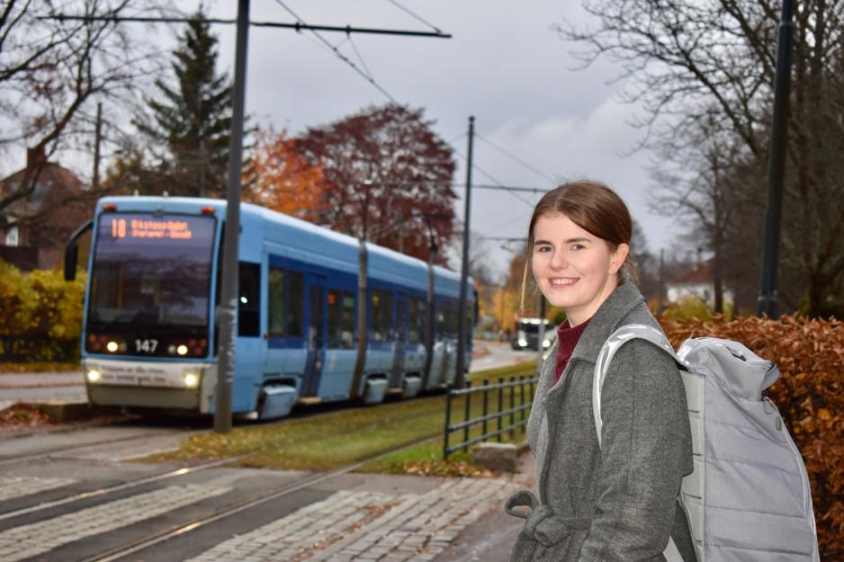 Ung kvinne med m&oslash;rkt h&aring;r og gr&aring; k&aring;pe venter p&aring; trikken. En trikk kj&oslash;rer i bakgrunnen. 
