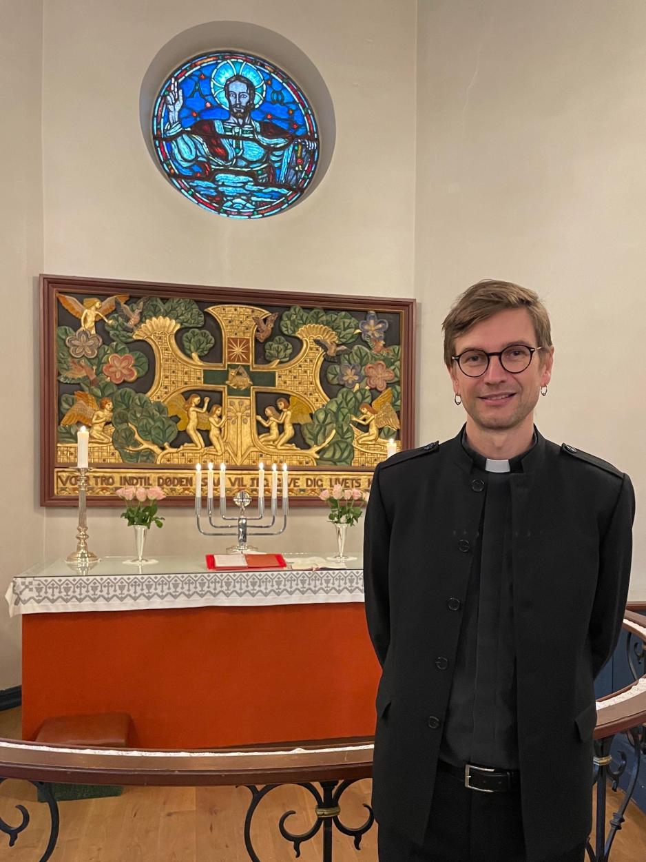 Bildet viser han som er prest i R&oslash;a og S&oslash;rkedalen menighet. Han st&aring;r foran alteret i S&oslash;rkedalen kirke, og p&aring; alteret er det lysestaker og rosa blomster.<