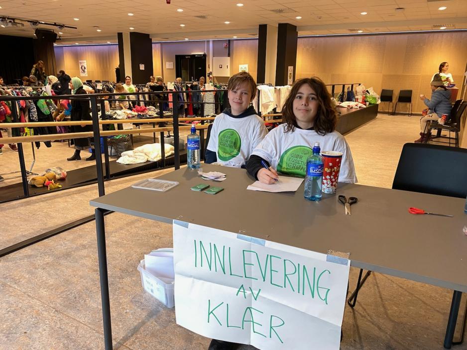 To unge gutter som sitter ved et bord. p&aring; bordet henger det et skilt som sier innlevering av kl&aelig;r. 