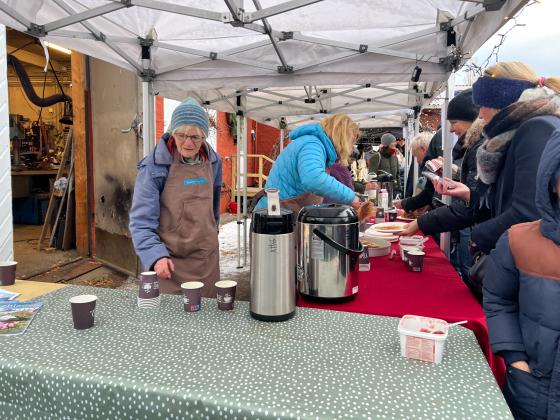 To kvinner fra venneforeningen botanisk hages venner som st&aring;r bak et bord. den bakerste gj&oslash;r klar en vaffel. kvinnen n&aelig;rmest kamera er p&aring; vei &aring; fylle p&aring; kaffe. utenfor bode er det folkerikt.