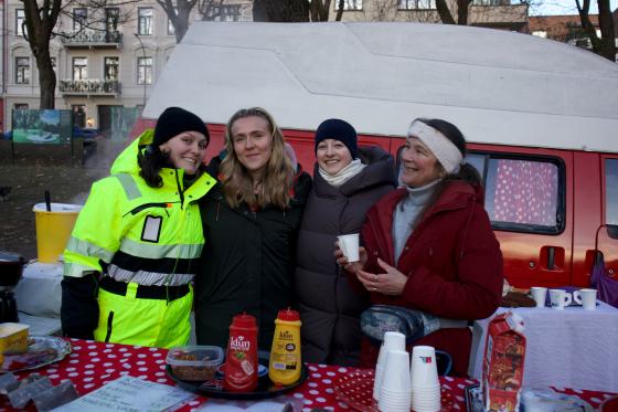 fire damer st&aring;r og smiler bak kaffebordet. I bakgrunnen er en r&oslash;d og hvit van 