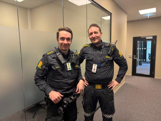 To politibetjenter som smiler inne p&aring; politistasjonen oppstilt ved siden av hverandre. 