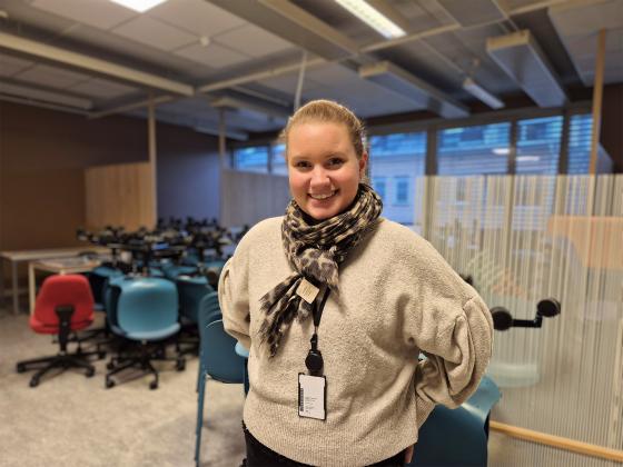 En dame st&aring;r foran et rom under oppussing fylt med stoler. Hun har h&aring;ret opp i en hestehale, og skjerf og genser p&aring;. Hun smiler og har hendene bak ryggen.