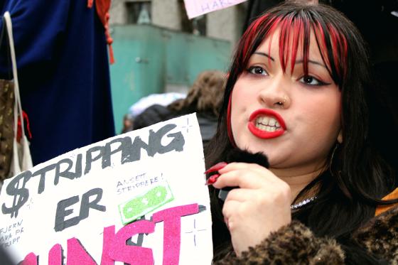 Stripper Alma Adore holder en plakat der det st&aring;r "Stripping er Kunst". Hun er midt i en setning og er derfor hard i fjeset og ser sint ut.