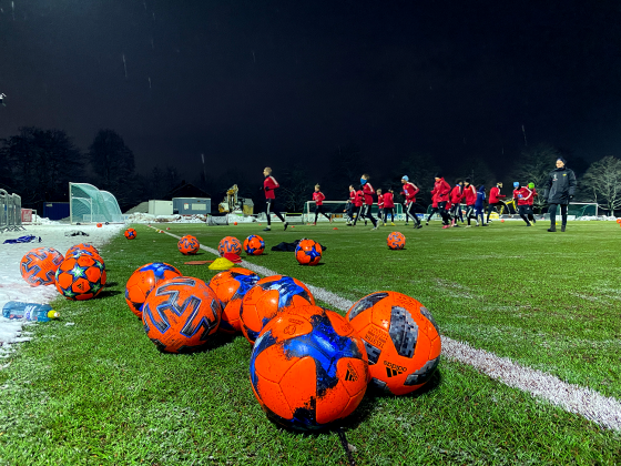 P&aring; fotballbane med spillere som trener i bakgrunn. Ser fotballer og en drikkeflaske.