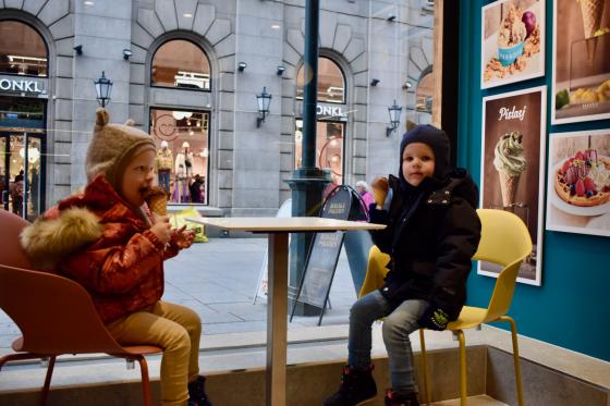To sm&aring; barn i parkdresser og luer sitter inne p&aring; en iskrembutikk og spiser is en kald novemberdag. Begge spiser en kule sjokoladeis i kjeks. Gjennom vinduet utenfor kan man se travle mennesker p&aring; Karl Johan. 