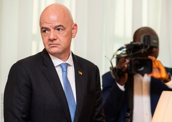 Bilde av Gianni Infantino i en svart dress med bl&aring;tt slips og en kameramann i bakgrunn.