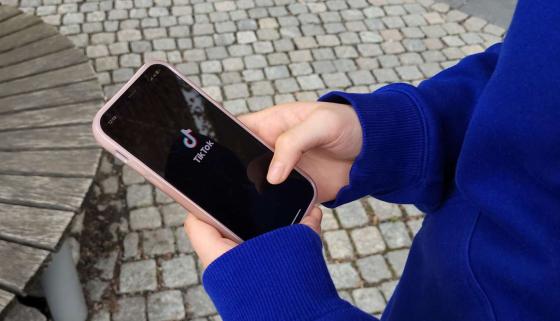 Bildet viser en jente som ser p&aring; telefonen sin. Hun har TikTok oppe og logoen til TikTok vises p&aring; skjermen. Jenta har p&aring; seg bl&aring; genser og st&aring;r ute. 