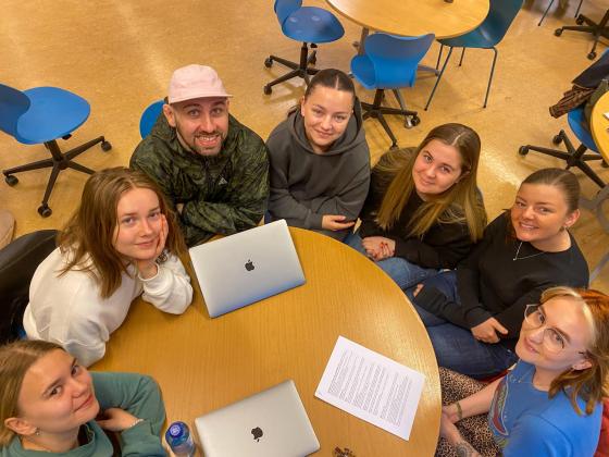sju studenter sitter rundt et rundt bor og ser opp
