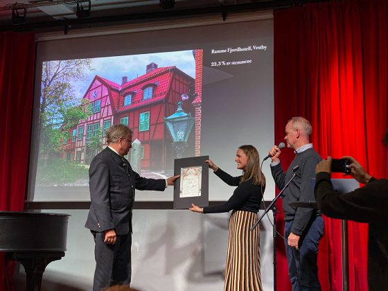 Bilde av tre folk som st&aring;r p&aring; en scene. Mannen f&aring;r en pris (hedersprisen). I bakgrunnen er det en powerpoint med bilde av Ramme Fjorhotell som ligger i Vestby.