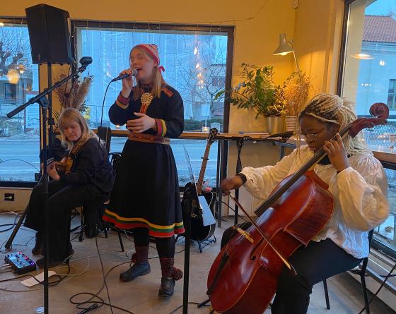En samisk jente st&aring;r og synger inn i en mikrofon med band rundt seg