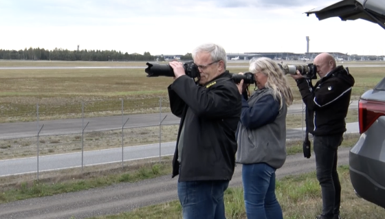 Tre flyspottere med kameraet mot ansiktet i h&oslash;yre hj&oslash;rnet av bildet. Rullebane p&aring; Gardermoen i bakgrunnen