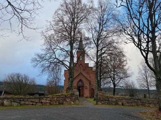 Bildet viser S&oslash;rkedalen kirke i Oslo. Kirken er laget av brune murstein, og har et h&oslash;yt kirkespir. Den st&aring;r litt tildekket av nakne l&oslash;vtr&aelig;r. 