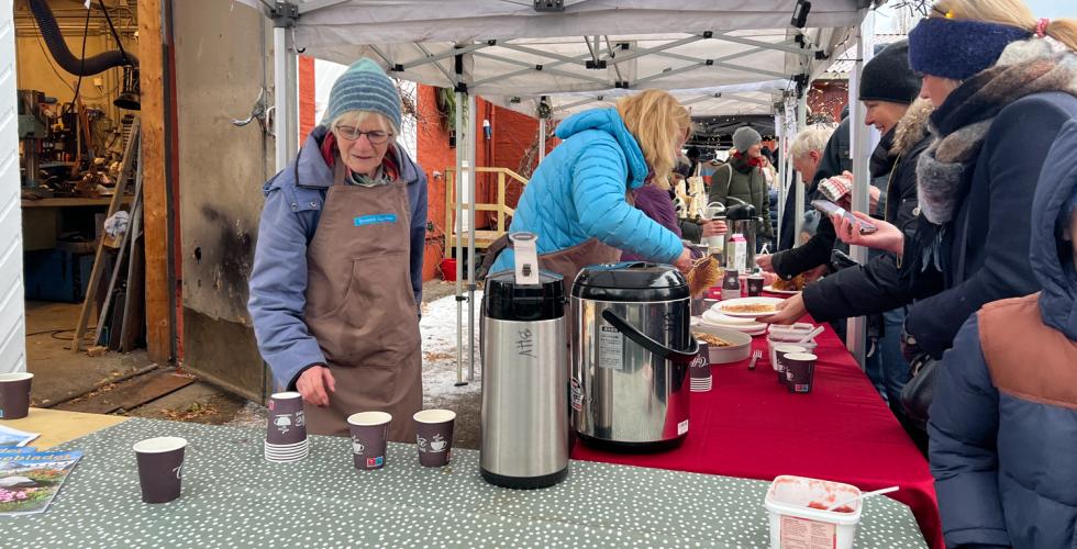 To kvinner fra venneforeningen botanisk hages venner som st&aring;r bak et bord. den bakerste gj&oslash;r klar en vaffel. kvinnen n&aelig;rmest kamera er p&aring; vei &aring; fylle p&aring; kaffe. utenfor bode er det folkerikt.