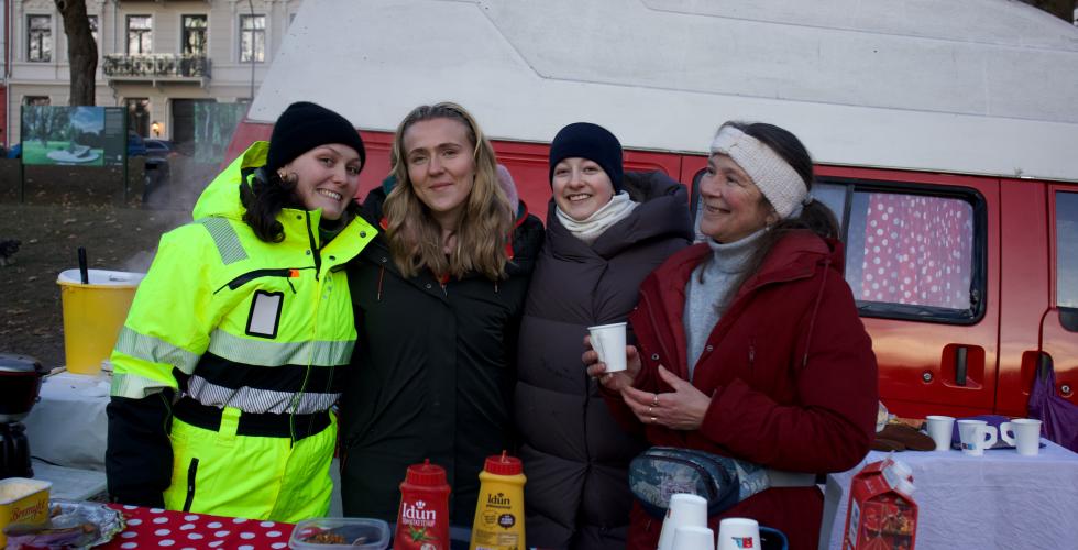 fire damer st&aring;r og smiler bak kaffebordet. I bakgrunnen er en r&oslash;d og hvit van 