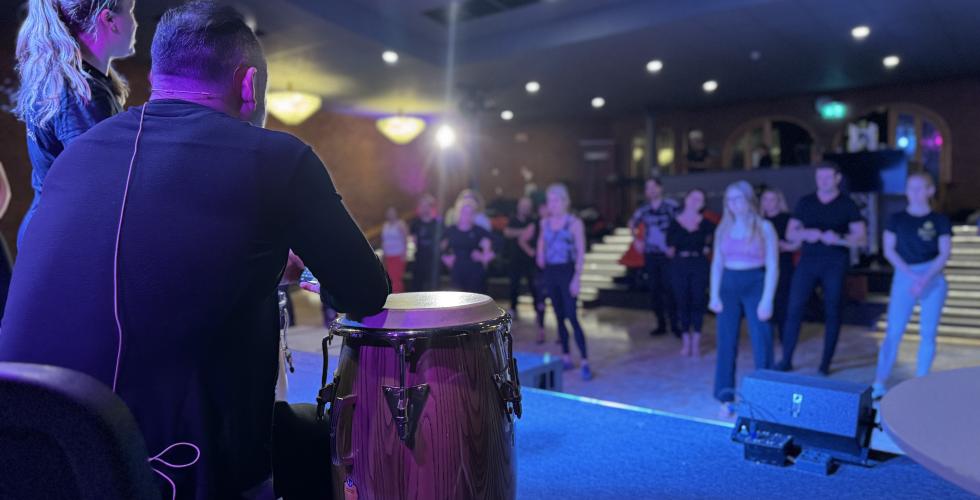 Bilde tatt fra scenen. I forgrunn sitter en instrukt&oslash;r og spiller trommer, mens en annen viser dans. I bakgrunnen st&aring;r en mengde elever som er p&aring; nybegynnerkurs i salsa. 
