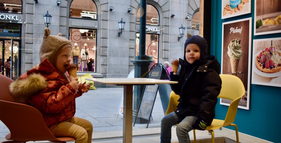 To sm&aring; barn i parkdresser og luer sitter inne p&aring; en iskrembutikk og spiser is en kald novemberdag. Begge spiser en kule sjokoladeis i kjeks. Gjennom vinduet utenfor kan man se travle mennesker p&aring; Karl Johan. 