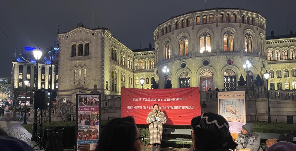 Bildet viser Khansa Ali som st&aring;r foran Stortinget og holder en appell. Bak henne henger det et r&oslash;dt banner hvor det st&aring;r "Beskytt voldsutsatte flyktningkvinner - fjern kravet om 5 &aring;rs botid for permanent opphold".