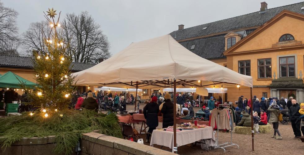 Bildet viser julemarkedet p&aring; Bogstad G&aring;rd. Man kan se et juletre med lys, et hvitt telt som holder regnet unna noen som selger produkter, og mange folk i bakgrunnen. 